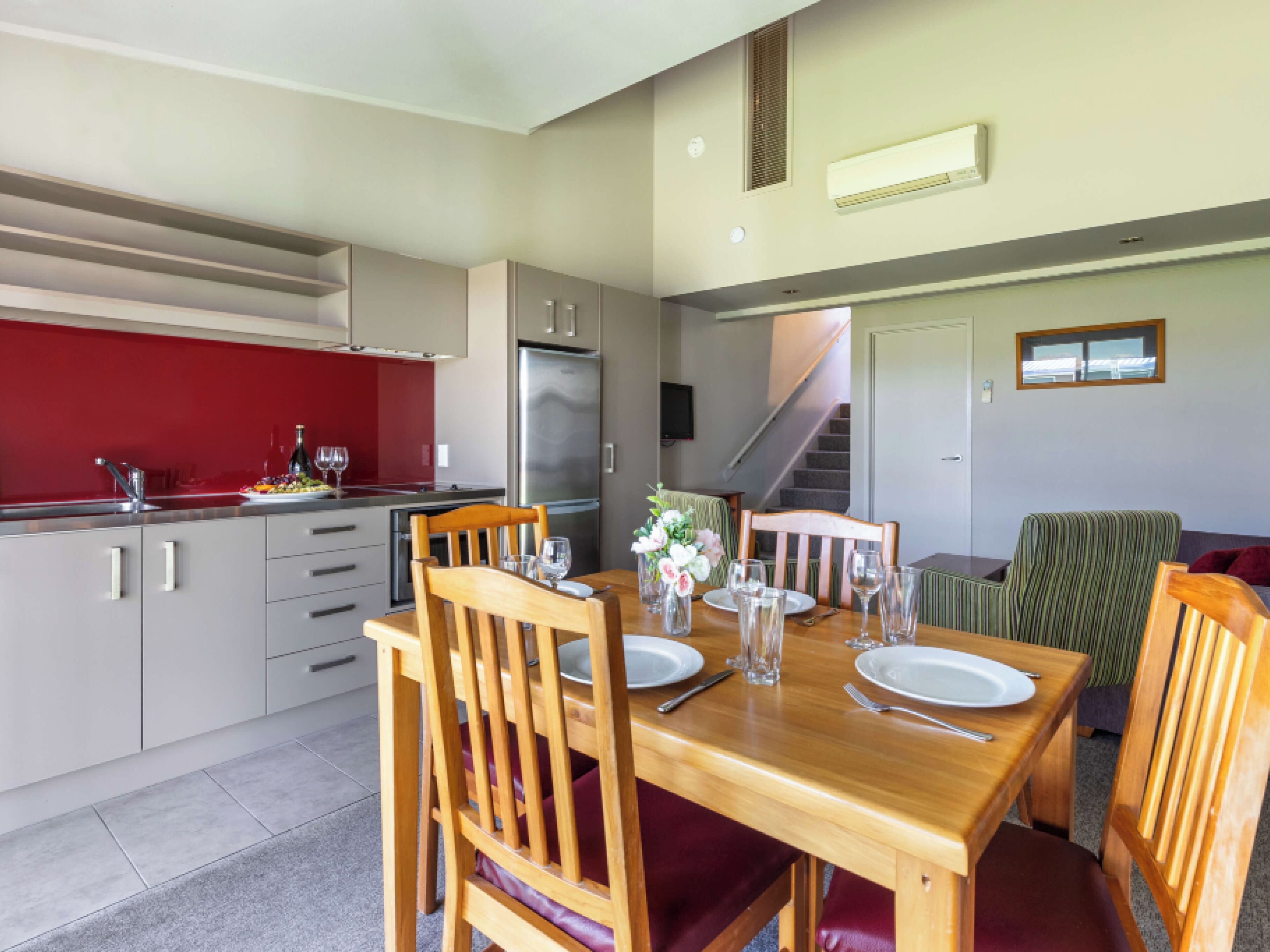 A modern kitchen with a red backsplash, stainless steel appliances, and a dining area with wooden furniture and a staircase leading to another level.