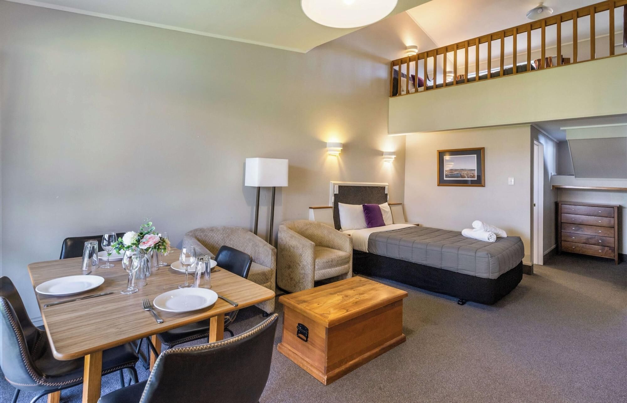 A hotel room with a dining table set for two, a sofa, a bed, a chest of drawers, and a staircase to an upper level.
