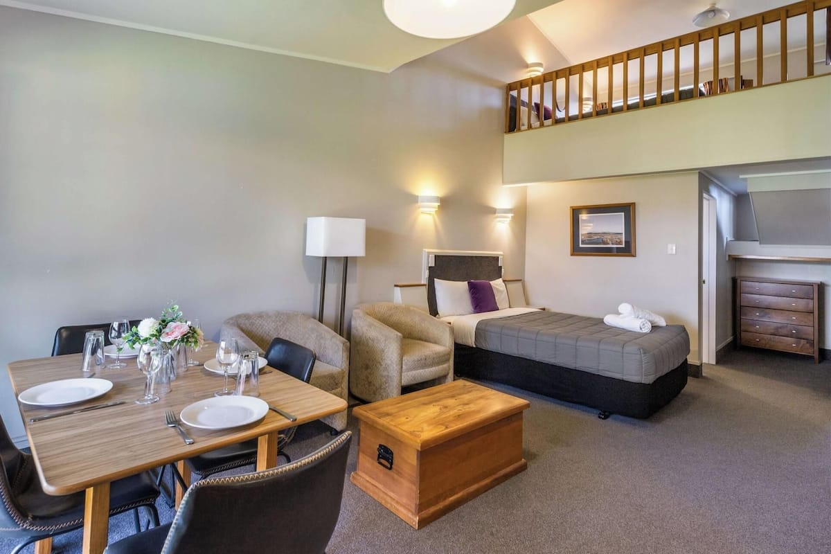 A hotel room with a dining table set for two, a sofa, a bed, a chest of drawers, and a staircase to an upper level.