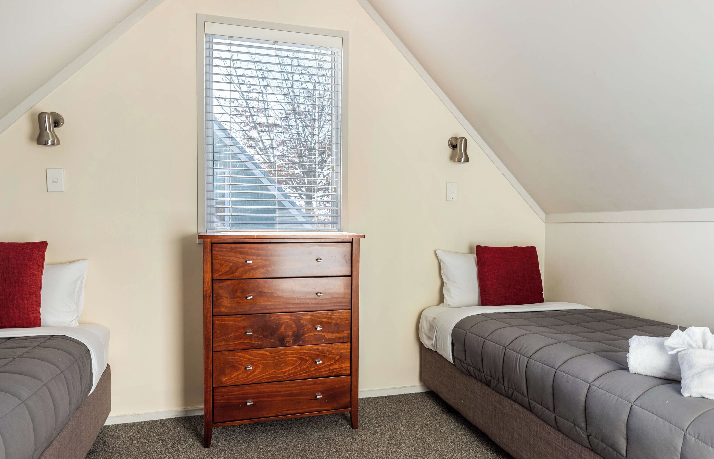 Ein Schlafzimmer mit schrägem Dach, einer Holzkommode, zwei Betten mit grauer Bettwäsche und einem Fenster mit Jalousien.