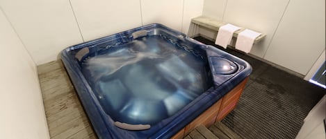 A hot tub in a room with white walls, a wooden floor, and a small bench with towels.