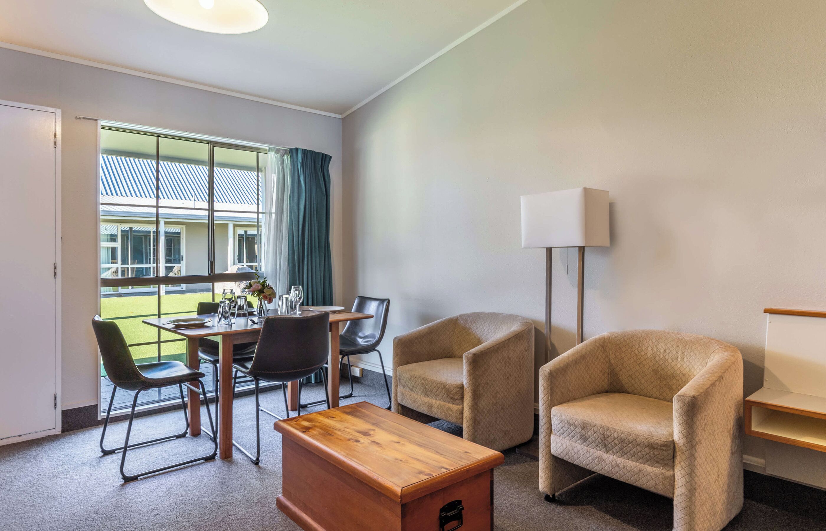 Habitación de hotel con comedor, dos sillones, una mesa de madera, una lámpara y vista a un edificio a través de una ventana.
