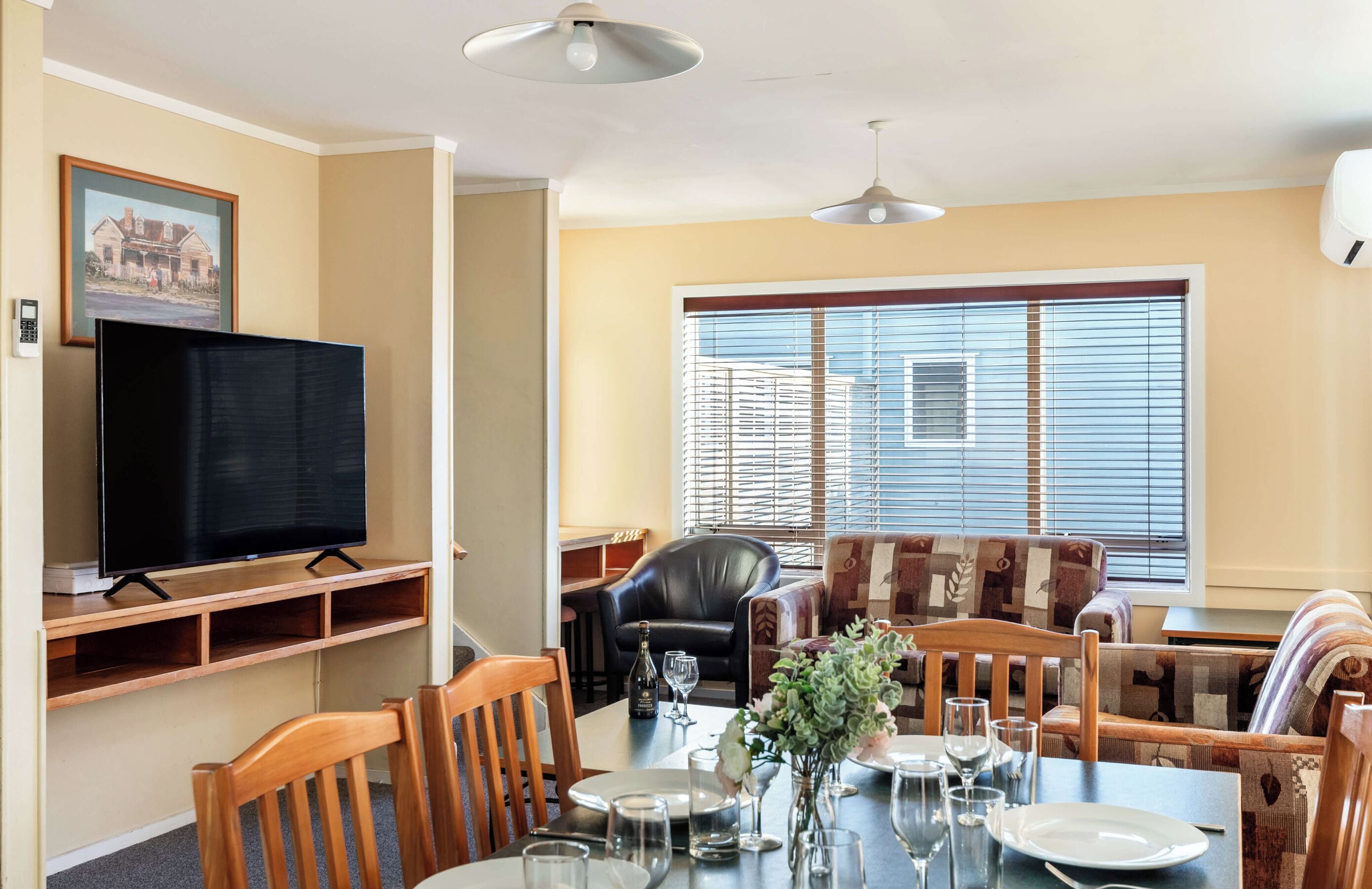 Un comedor con una mesa dispuesta para dos personas, un televisor sobre un mueble bajo y un sofá con un cojín estampado.