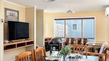 Un comedor con una mesa dispuesta para dos personas, un televisor sobre un mueble bajo y un sofá con un cojín estampado.