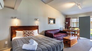A hotel room with a large bed, a wooden headboard, a red leather sofa, a wooden coffee table, and a dining area with a table and chairs. - Turangi Leisure Lodge (Turangi)
