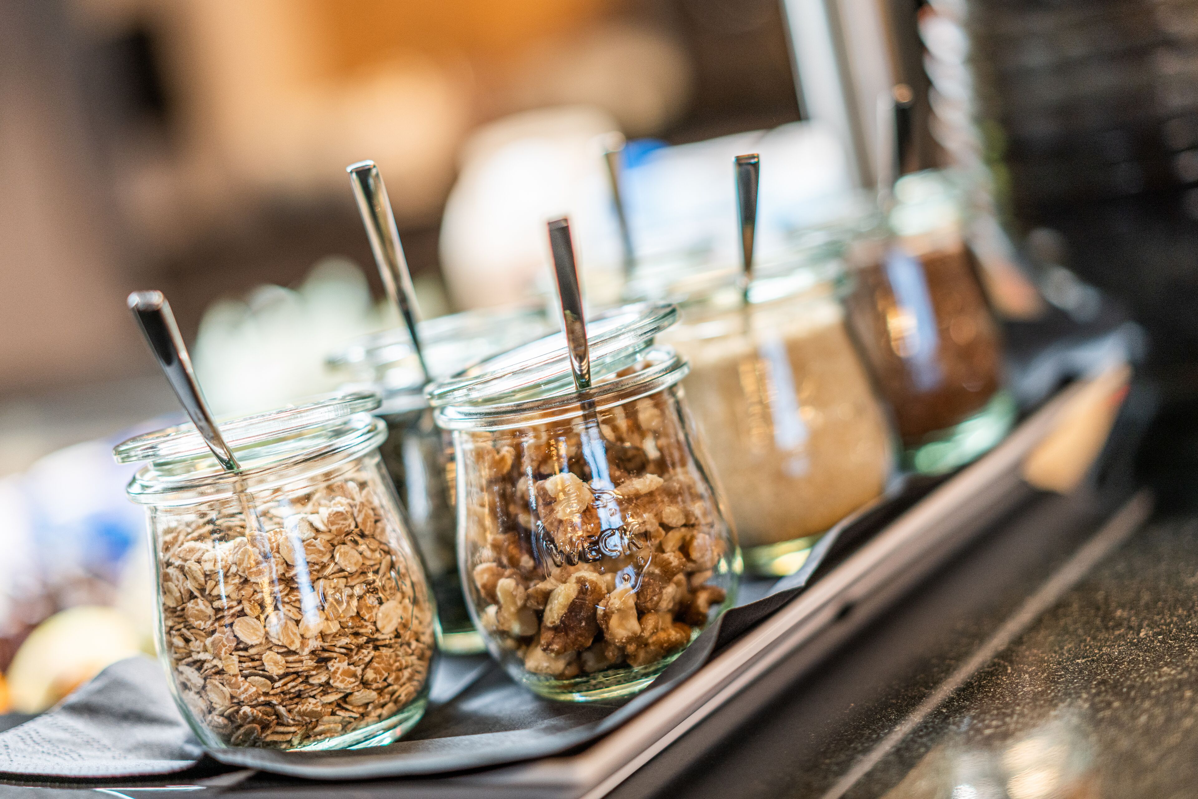 A row of glass jars with metal spoons, containing different food items.