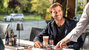 Un uomo seduto a un tavolino di un caffè all'aperto, con un bicchiere e degli accompagnamenti.