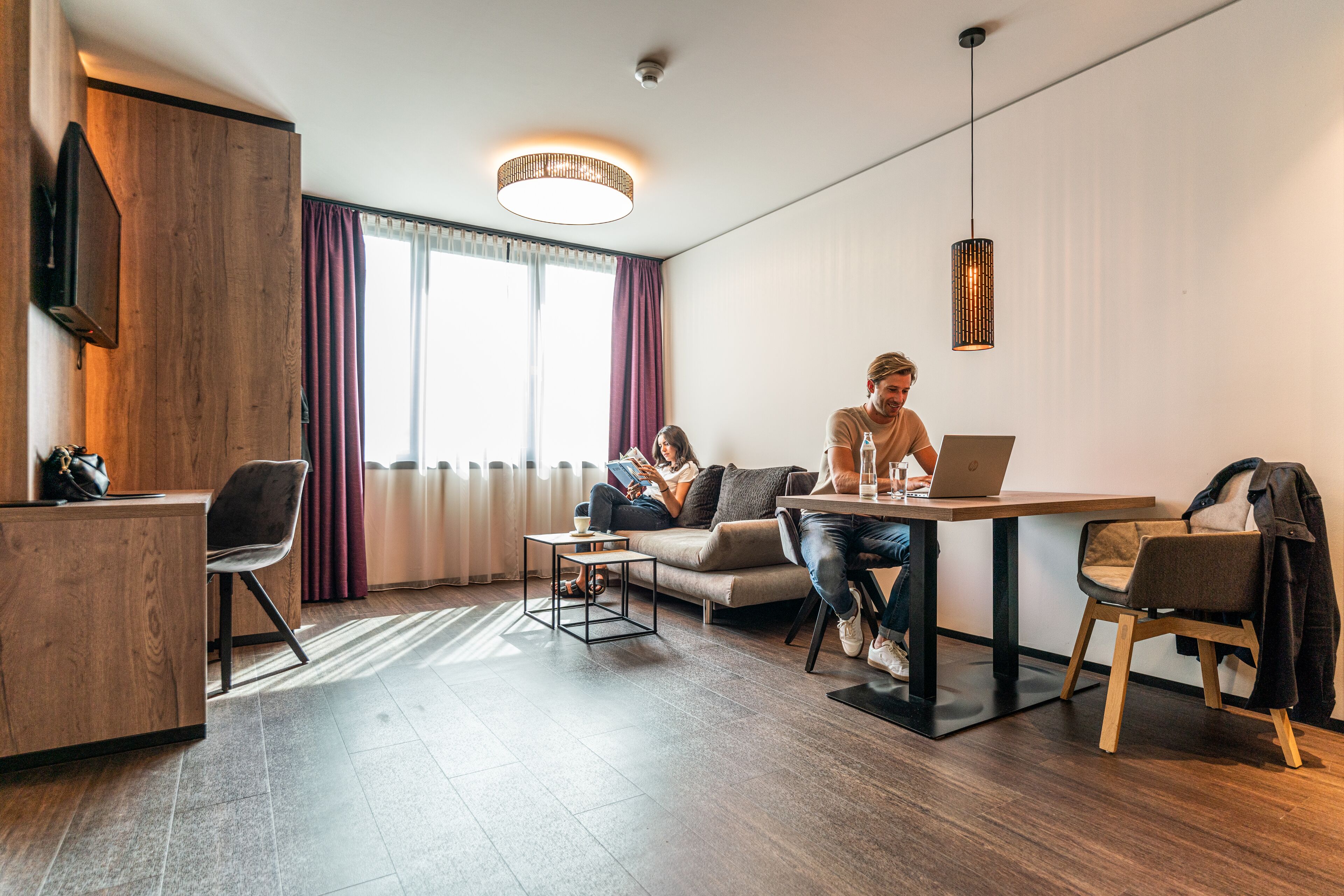 A modern hotel room with a wooden desk, a sofa, a dining table, and a large window with curtains.