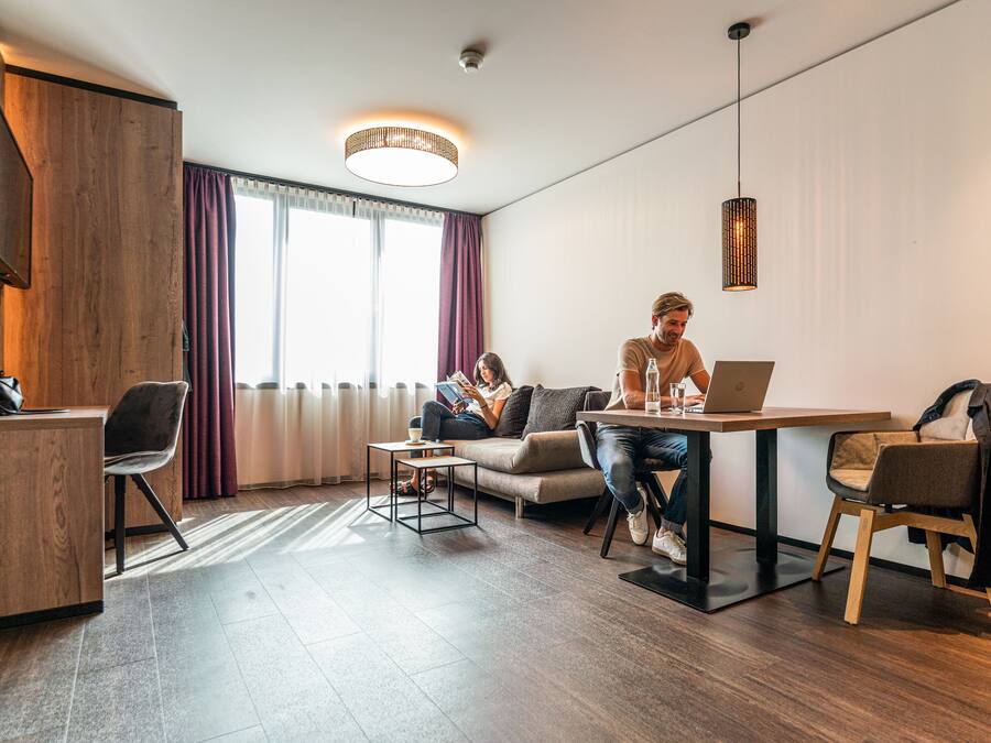 A modern hotel room with a wooden desk, a sofa, a dining table, and a large window with curtains.