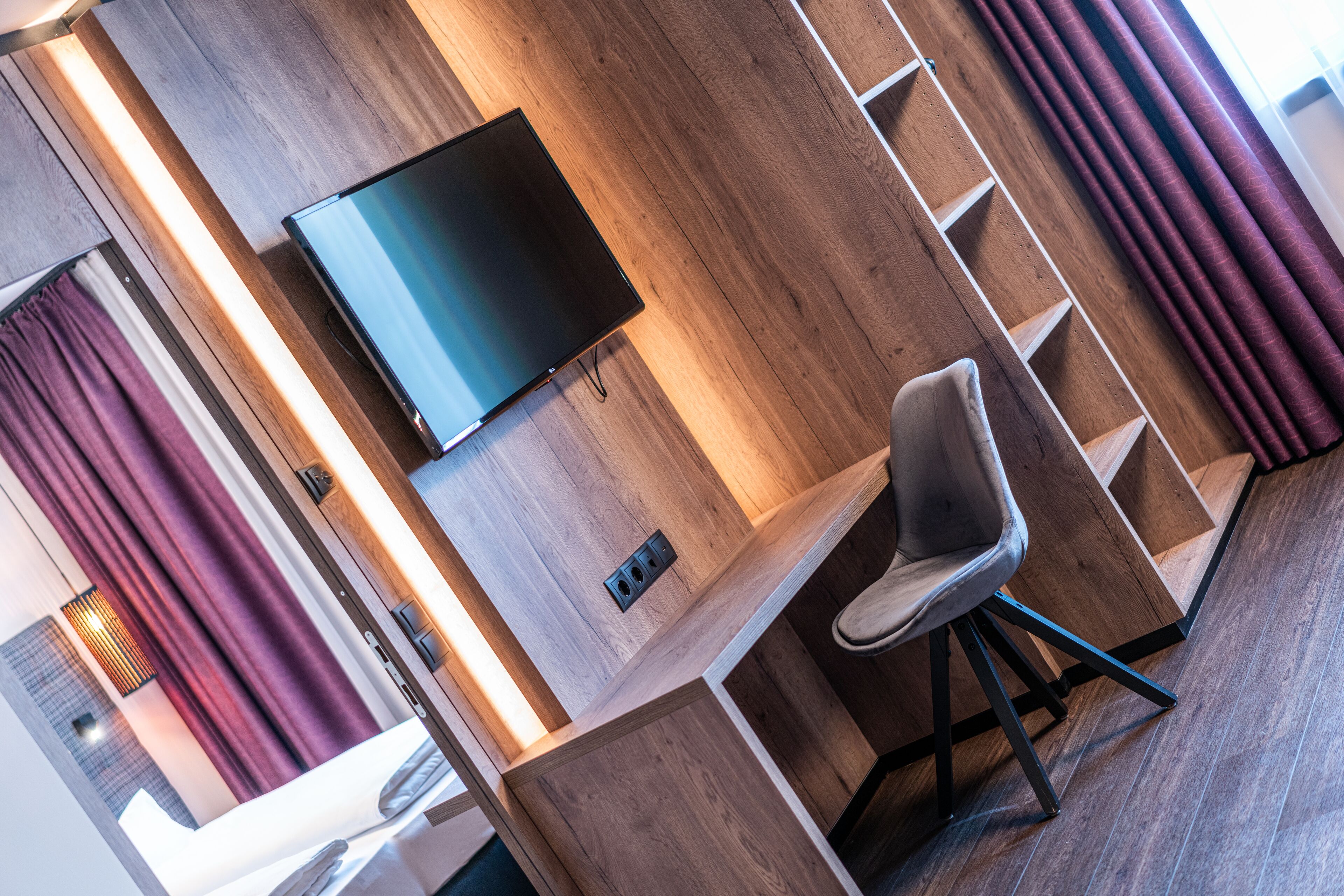 A modern hotel room with a wooden floor, a flat-screen TV mounted on the wall, a comfortable chair, and a built-in desk.