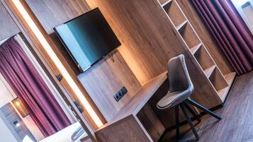 A modern hotel room with a wooden floor, a flat-screen TV mounted on the wall, a comfortable chair, and a built-in desk.