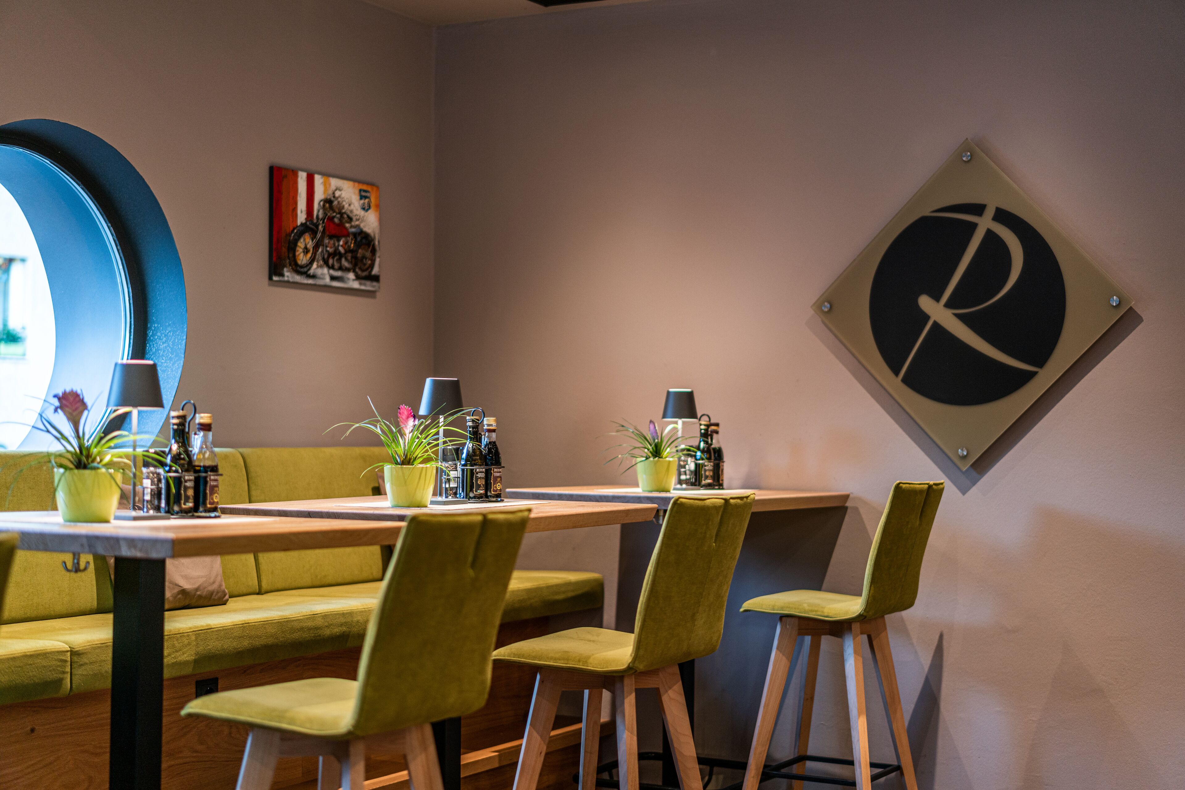 A dining area with a wooden table, green chairs, and a sofa. There are potted plants on the table and a motorcycle painting on the wall.