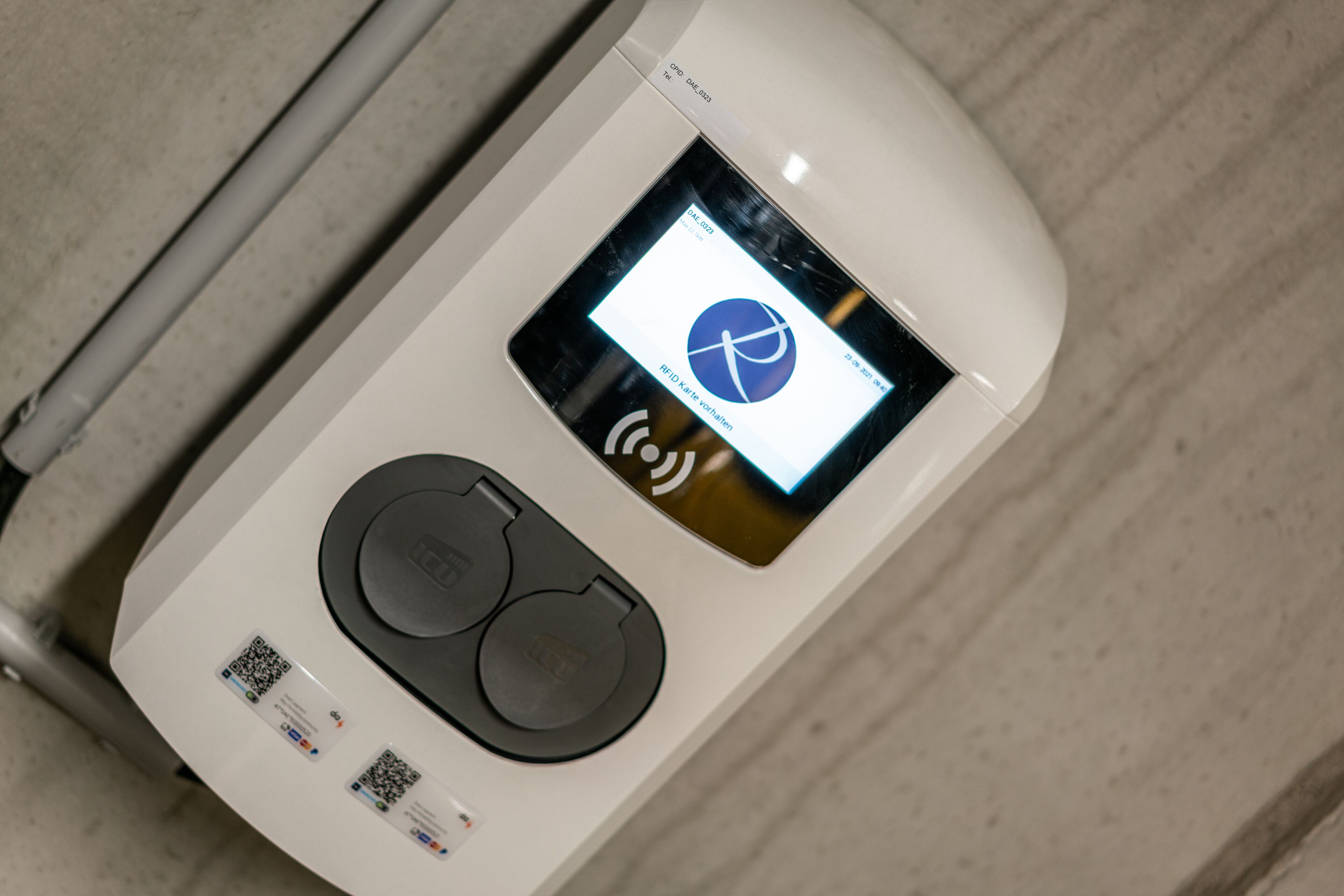 Un dispositivo blanco y moderno con una pantalla que muestra un logo, texto, dos botones circulares y códigos QR.