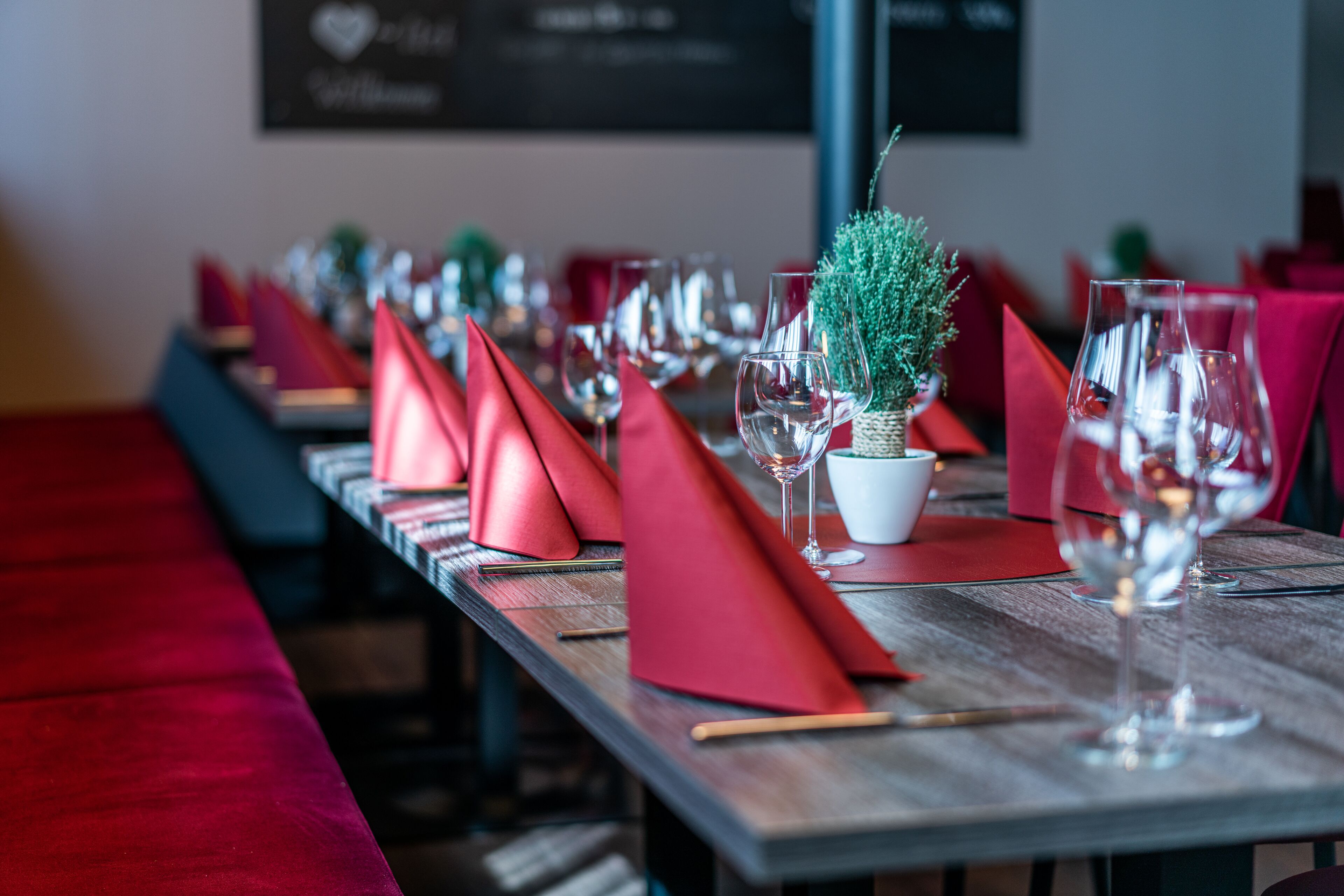 A restaurant setting with a long wooden table set with red napkins, wine glasses, and a small potted plant.