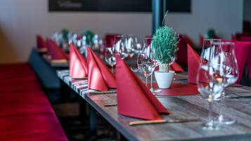 Ambiente de restaurante com uma mesa de madeira comprida, disposta com guardanapos vermelhos, copos de vinho e uma pequena planta em vaso.