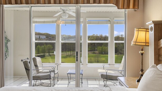 A bedroom with a large window, a ceiling fan, and a lamp. - Walk-in Golf Condo with Course View from All-Season Sunroom (Branson)