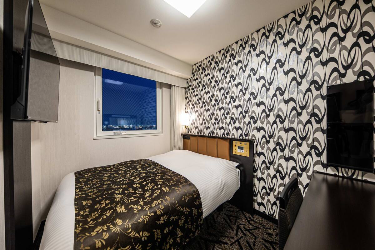 A hotel room with a bed, a desk, a TV, a window with a city view, and patterned wallpaper.