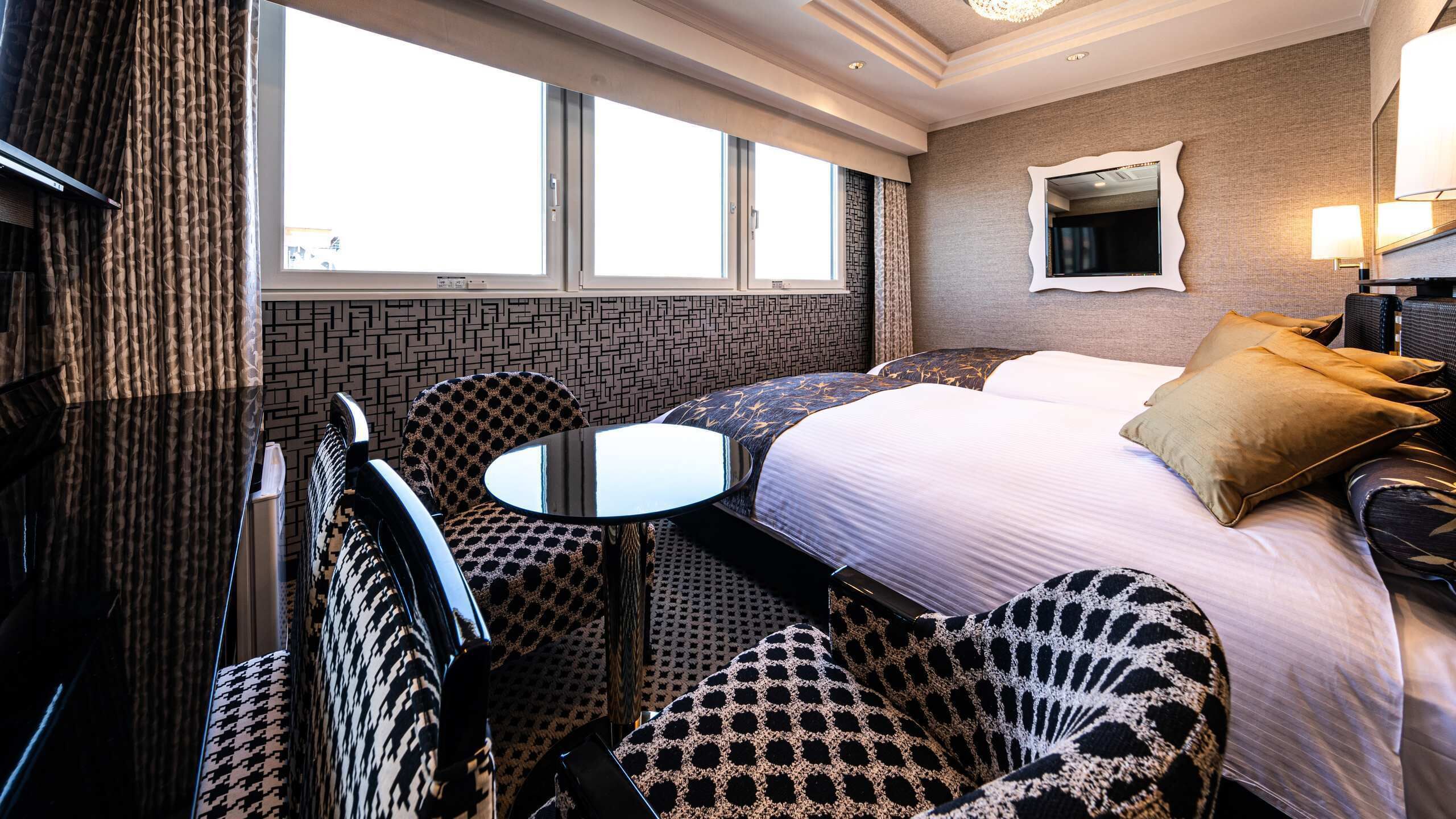 Quarto de hotel com cama, duas poltronas, uma mesinha, um espelho e uma janela.