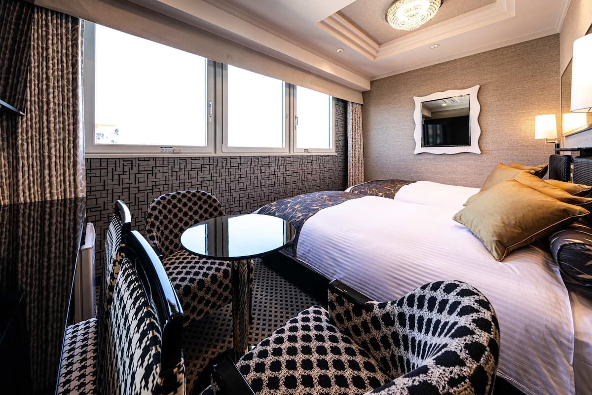 A hotel room with a bed, two chairs, a small table, a mirror, and a window with curtains.