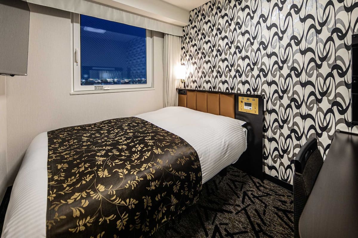 A hotel room with a bed, a window, a patterned wall, and a television.