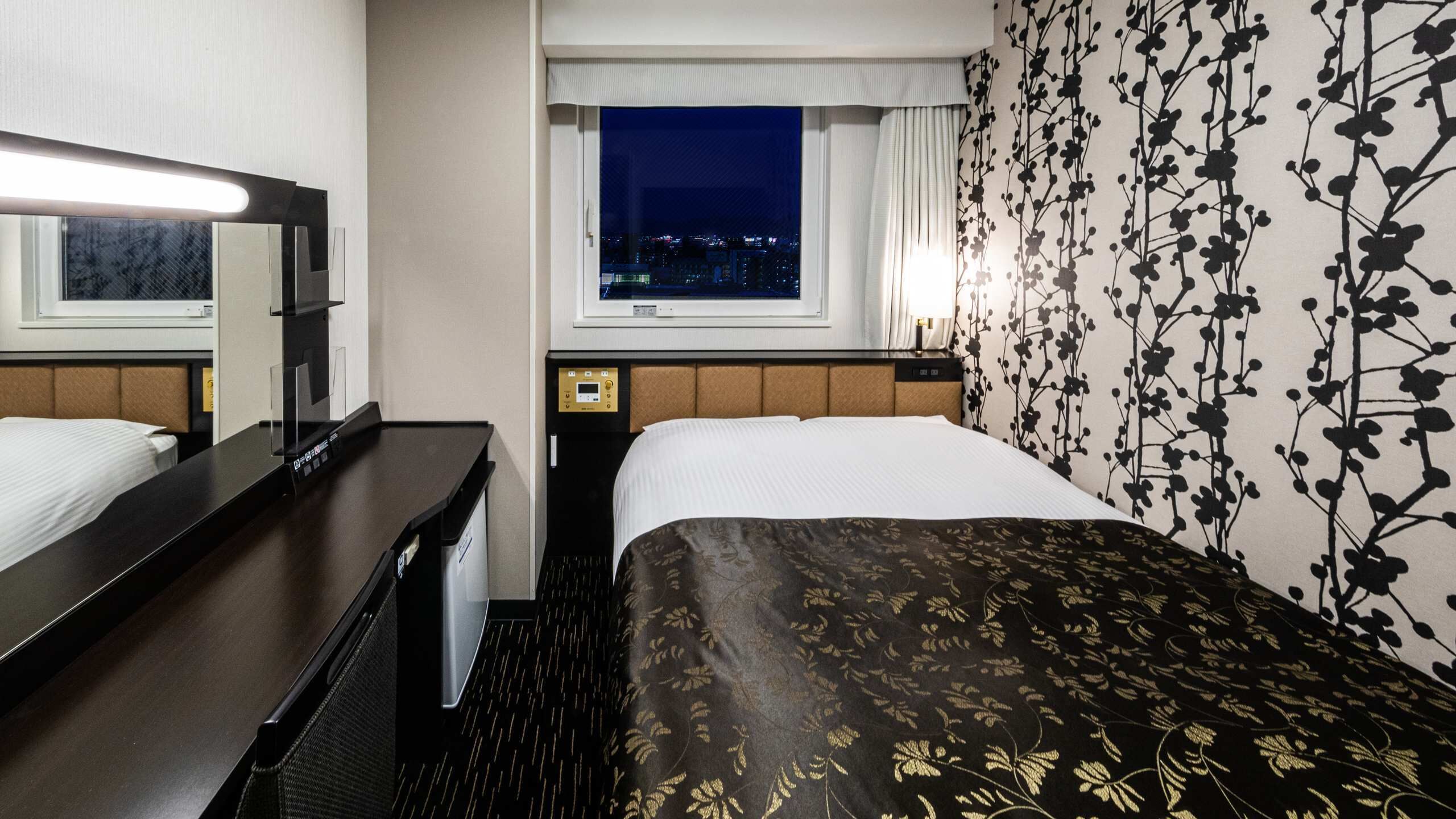 Quarto de hotel com cama, escrivaninha, espelho, janela com vista noturna e parede com padrão floral.