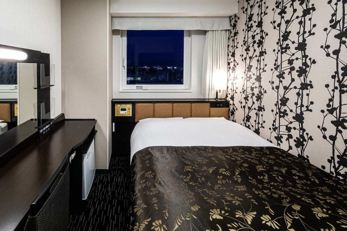 A hotel room with a bed, a desk, a mirror, a window with a night view, and a wall with a floral pattern.