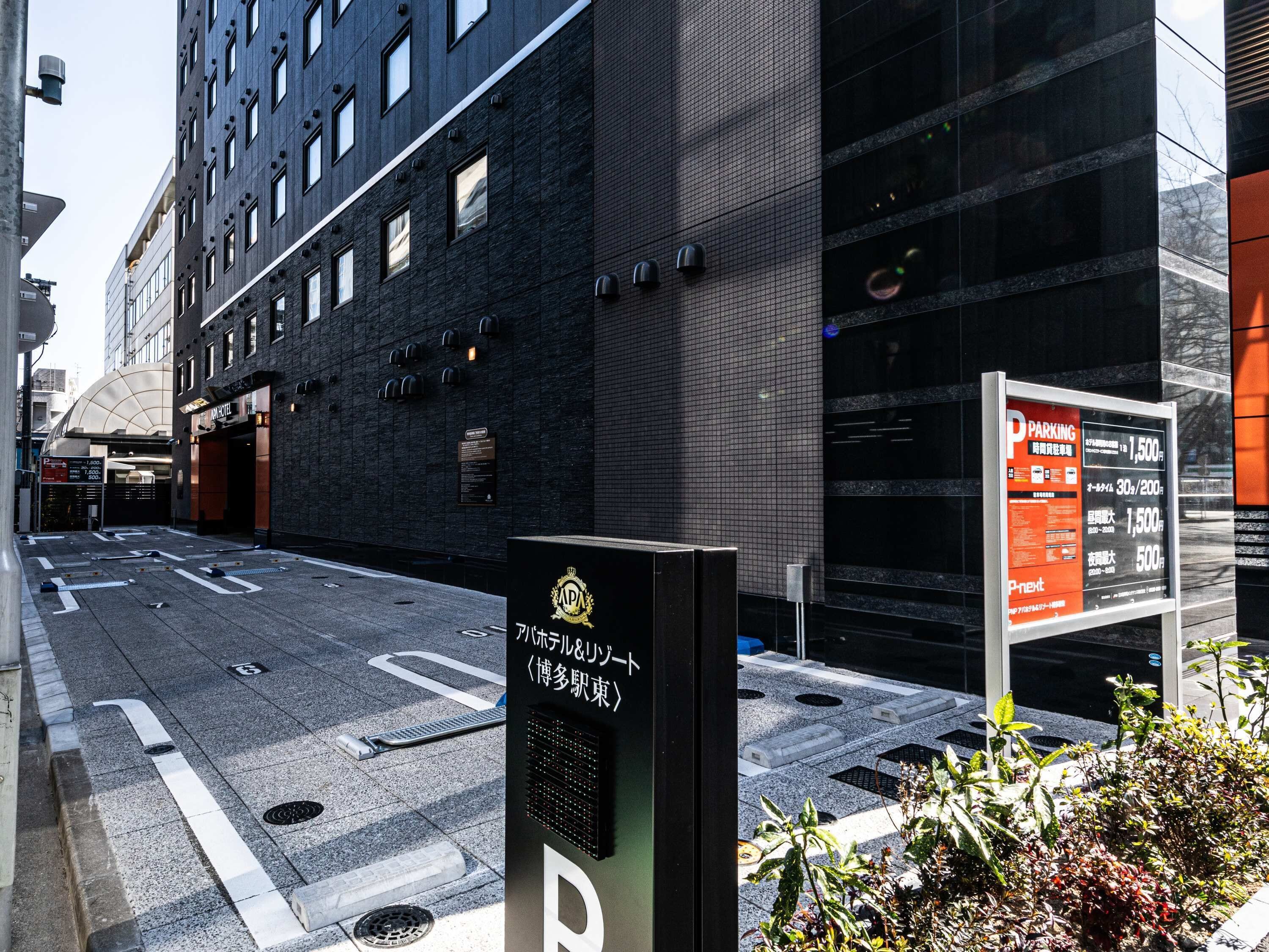Segno di parcheggio con informazioni sui prezzi in una lingua straniera, un edificio con una facciata moderna e un cartello con testo giapponese.