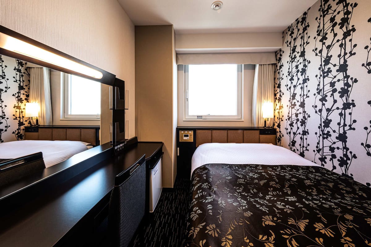 A hotel room with two beds, a large mirror, a desk, a window, and a wall with a floral pattern.