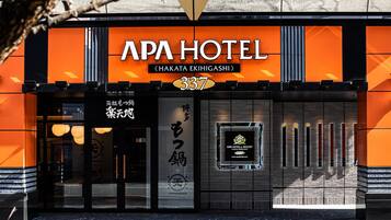 Exterior del APA Hotel con un llamativo frente naranja, una entrada amplia y un cartel que muestra el nombre y la dirección del hotel.