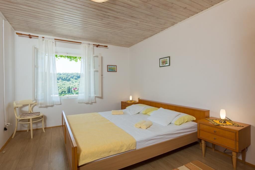 A bedroom with a wooden bed, a nightstand with a lamp, a chair, and a window with curtains.