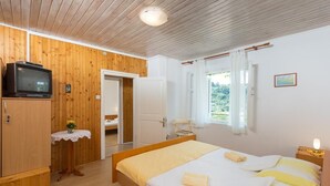 Quarto com teto de madeira, cama com roupa de cama branca e amarela, uma mesinha com um vaso e uma televisão sobre um armário de madeira.