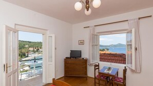 Quarto com cômoda de madeira, televisão, mesa de jantar com cadeiras, tapete vermelho e vista para o mar e colinas pela janela.