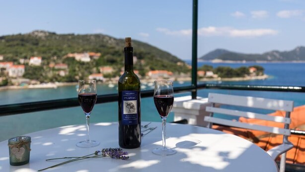 Uma mesa com uma garrafa de vinho Plavac, duas taças de vinho tinto e uma vela numa varanda com vista para a costa.