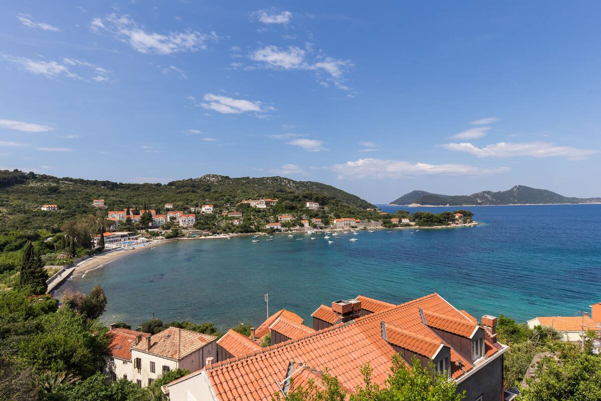 A coastal town with terracotta-roofed buildings, a sandy beach, and clear blue waters.
