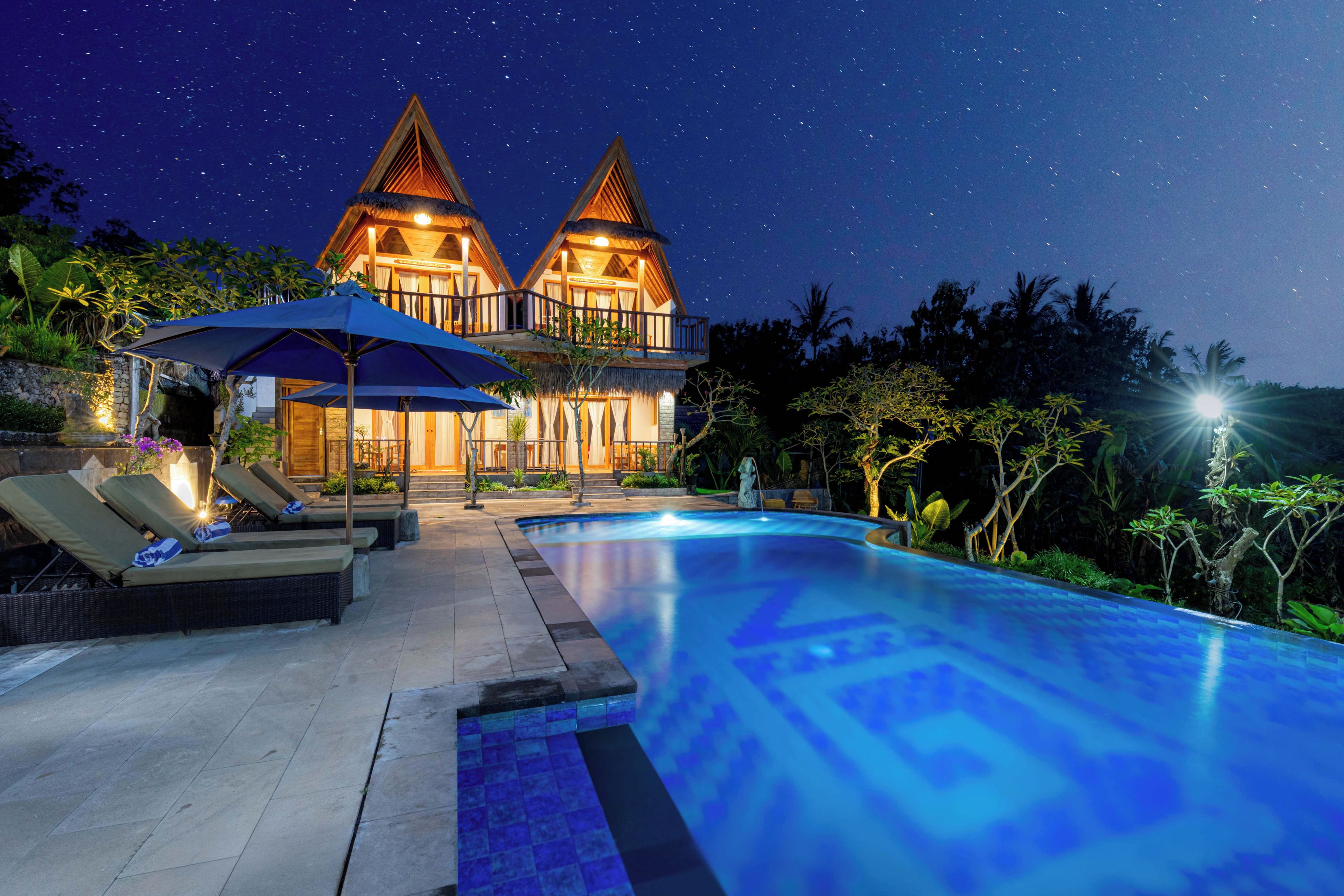 A two-story villa with a swimming pool, lounge chairs, and a thatched roof.