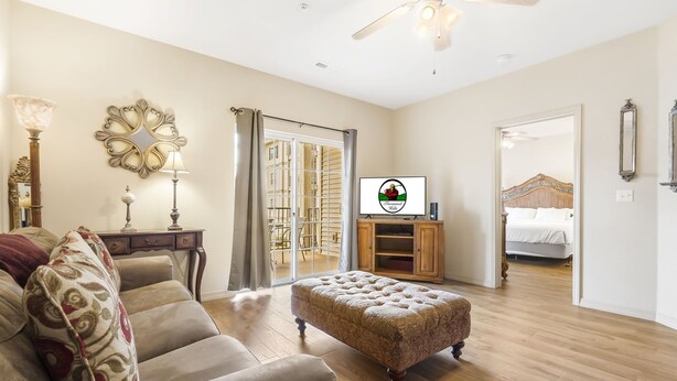 A spacious living room with a wooden floor, a large ottoman, a sofa, a television, and a dining area visible through a glass door. - Walk-In 2BR Branson Condo Near Strip | Sleeps 7 | Golf + Ticket Deals (Branson)