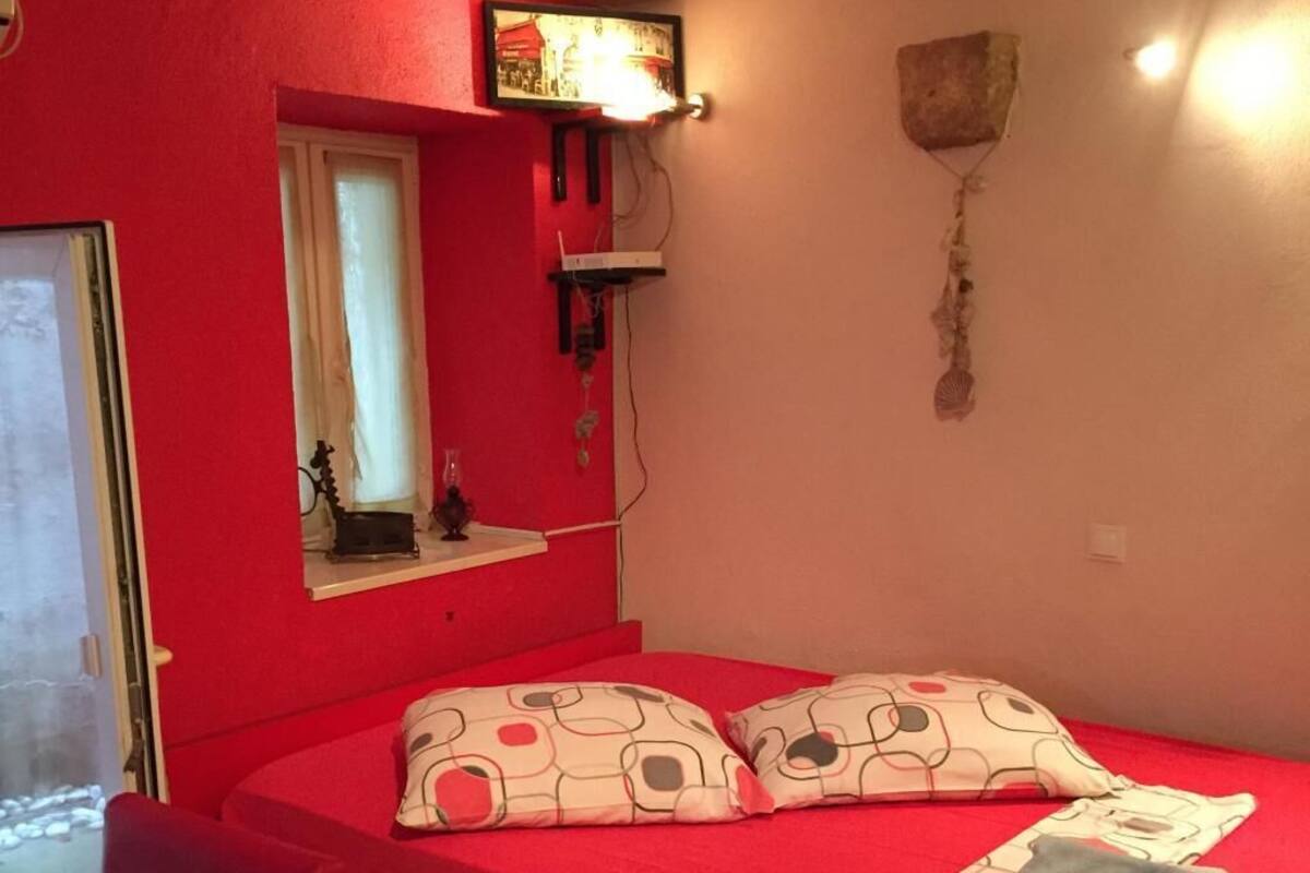 Una habitación con una cama roja, un ventanal pequeño, un estante en la pared y una puerta.