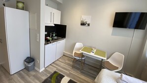 Une cuisine compacte avec des meubles blancs, un plan de travail noir et un petit évier. On y trouve un coin repas avec deux chaises et une table, une poubelle et une horloge au mur.