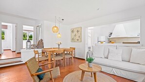 Una sala de estar moderna con un sofá blanco, una mesa de comedor de madera, sillas de mimbre, una mesa redonda de centro y una lámpara colgante.