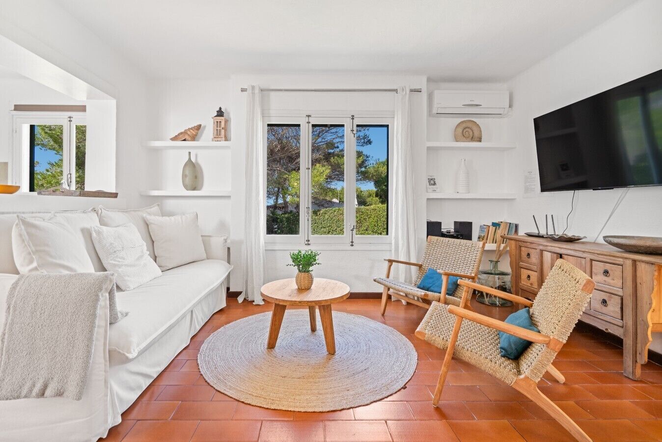 A modern living room with a white sofa, a round wooden table with a plant, a wicker chair, and a TV on the wall.