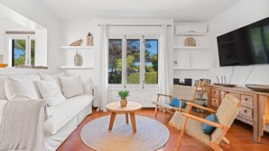 Una sala de estar moderna con un sofá blanco, una mesa redonda de madera con una planta, una silla de mimbre y un televisor en la pared.