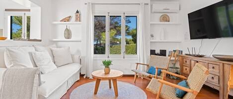 A modern living room with a white sofa, a round wooden table with a plant, a wicker chair, and a TV on the wall.
