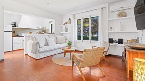 Un salón moderno con un sofá blanco, una mesa de centro redonda de madera, sillas de mimbre, una zona de cocina con armarios blancos y un televisor montado en la pared.