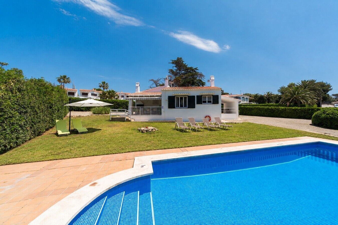 A poolside view of a house with a swimming pool, a covered patio, and a lawn with lounge chairs.