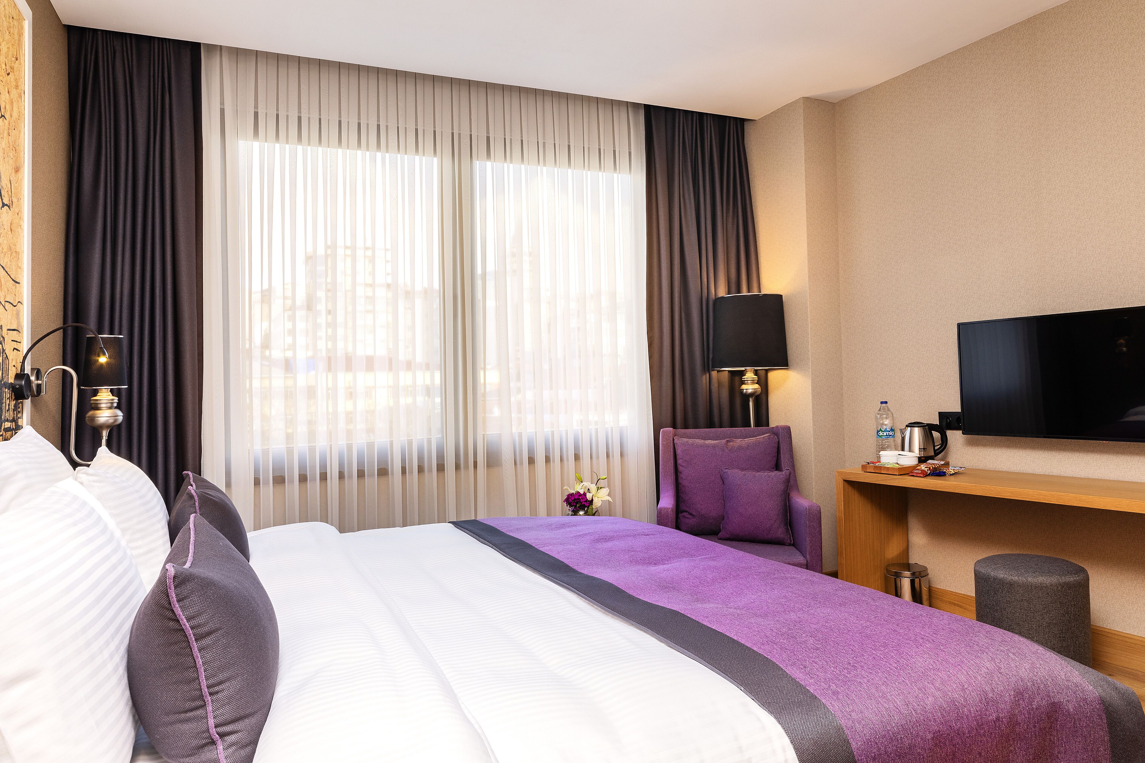A hotel room with a large bed, a TV, a desk with a lamp, and a window with sheer curtains.