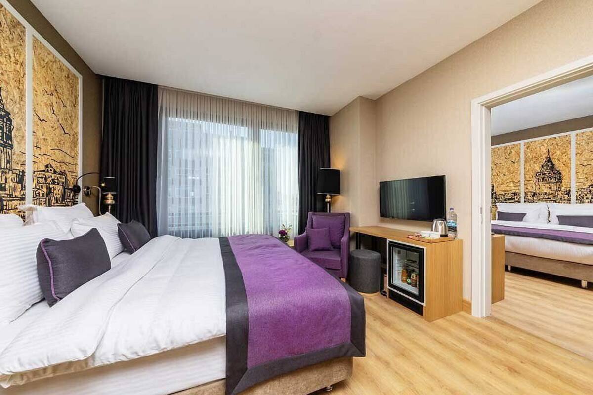 A hotel room with a large bed, a desk with a TV, a chair, and a minibar.