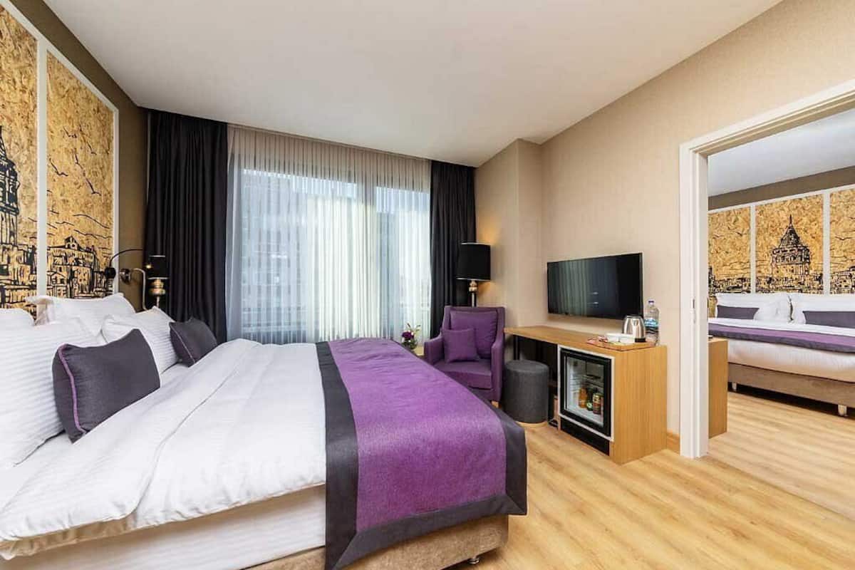 Ein Hotelzimmer mit einem großen Bett, einem Schreibtisch mit Fernseher, einem Sessel und einer Minibar.