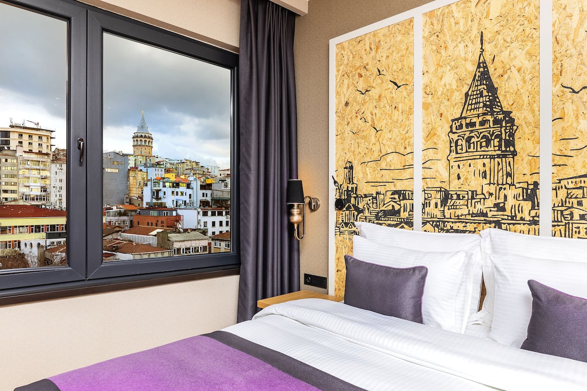 A hotel room with a bed, a view of a cityscape through a window, and a wall with a cityscape mural.