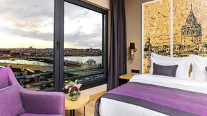 A hotel room with a large window offering a city view, a bed with purple bedding, a small round table with a vase of flowers, and a wall-mounted lamp. - The Halich Hotel Istanbul Karakoy (Istanbul)