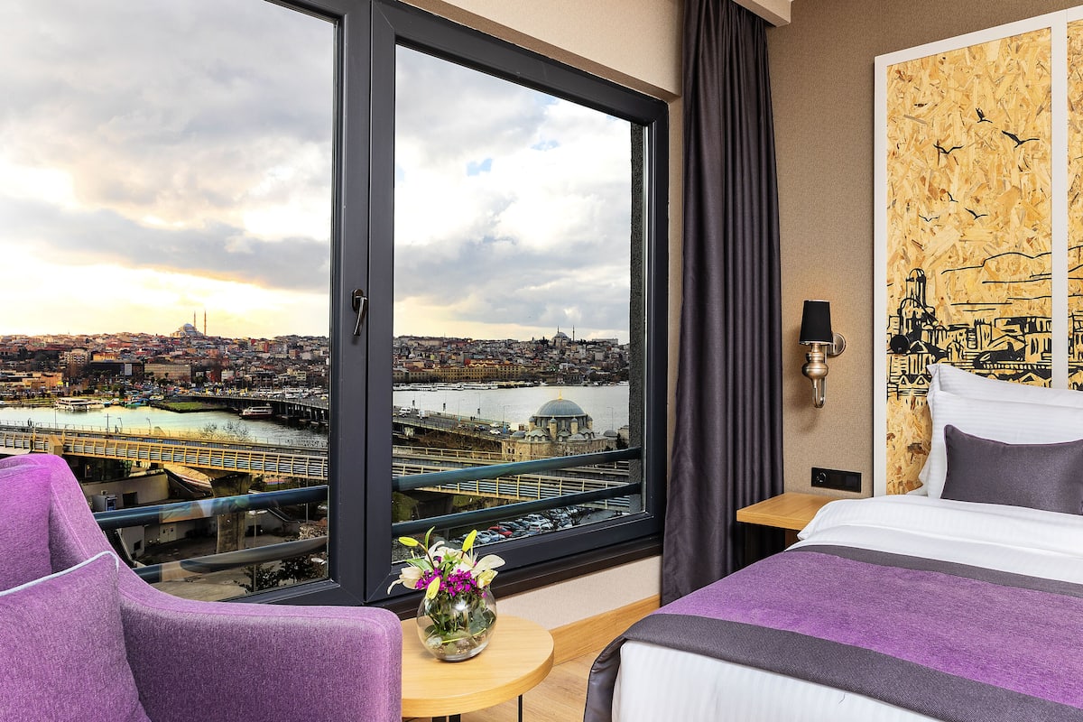 A hotel room with a large window offering a city view, a bed, a small table with a vase of flowers, and a wall-mounted lamp.